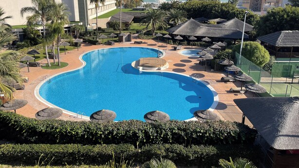 Una gran piscina de diseño curvo, rodeada de sillas de descanso y sombrillas, con vista al océano y edificios al fondo.