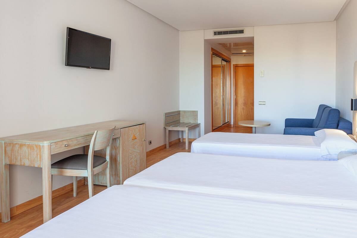 Ein Hotelzimmer mit zwei Betten, einem Schreibtisch, einem Sessel, einem Fernseher und einem kleinen Tisch.
