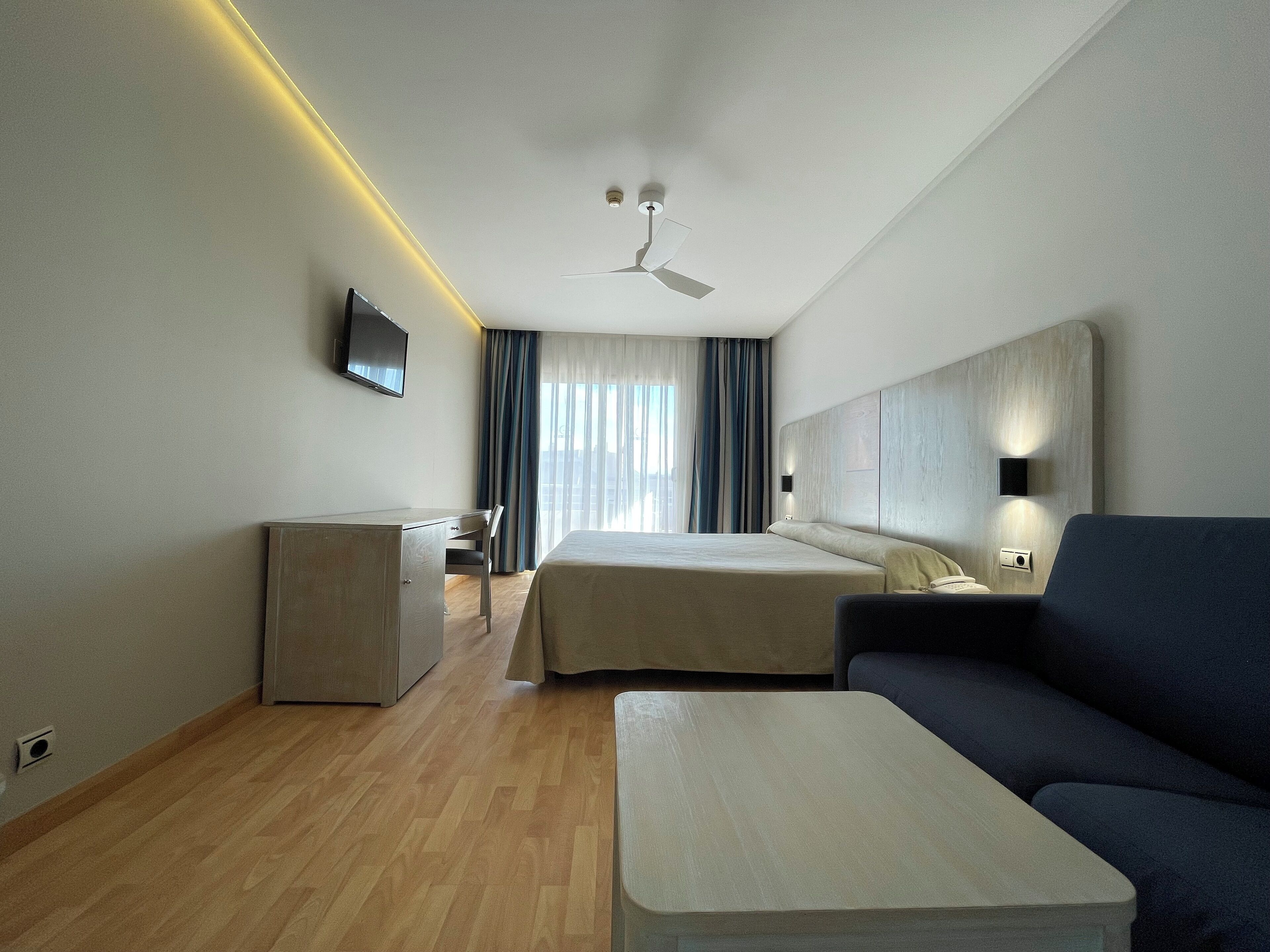 A modern hotel room with a bed, a desk, a chair, a sofa, and a television.