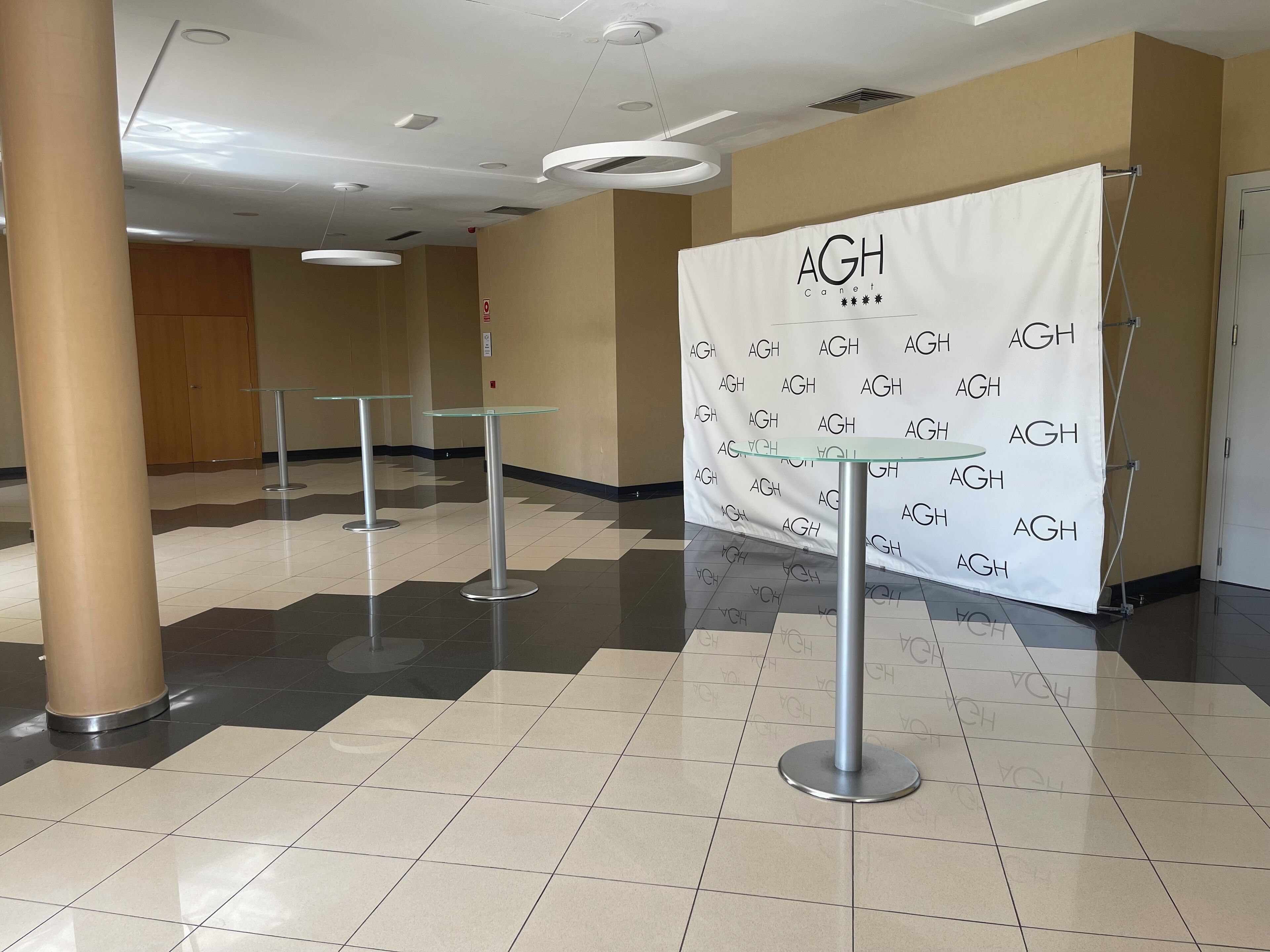 Un vestíbulo con un cartel blanco que repite “AGH Canet”, mesas de vidrio y suelo de baldosas.