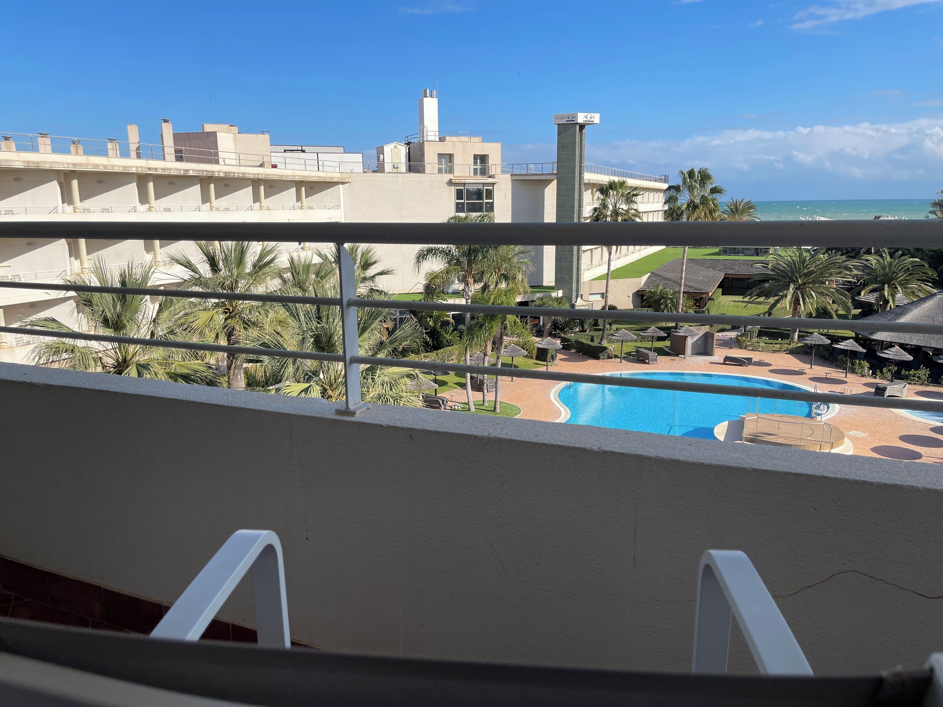 A view from a balcony overlooking a swimming pool, palm trees, and a hotel building.