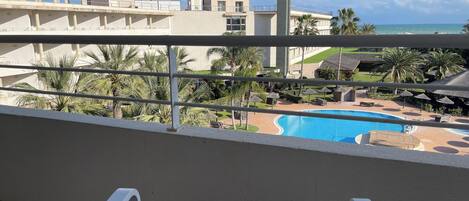 A view from a balcony overlooking a swimming pool, palm trees, and a hotel building.