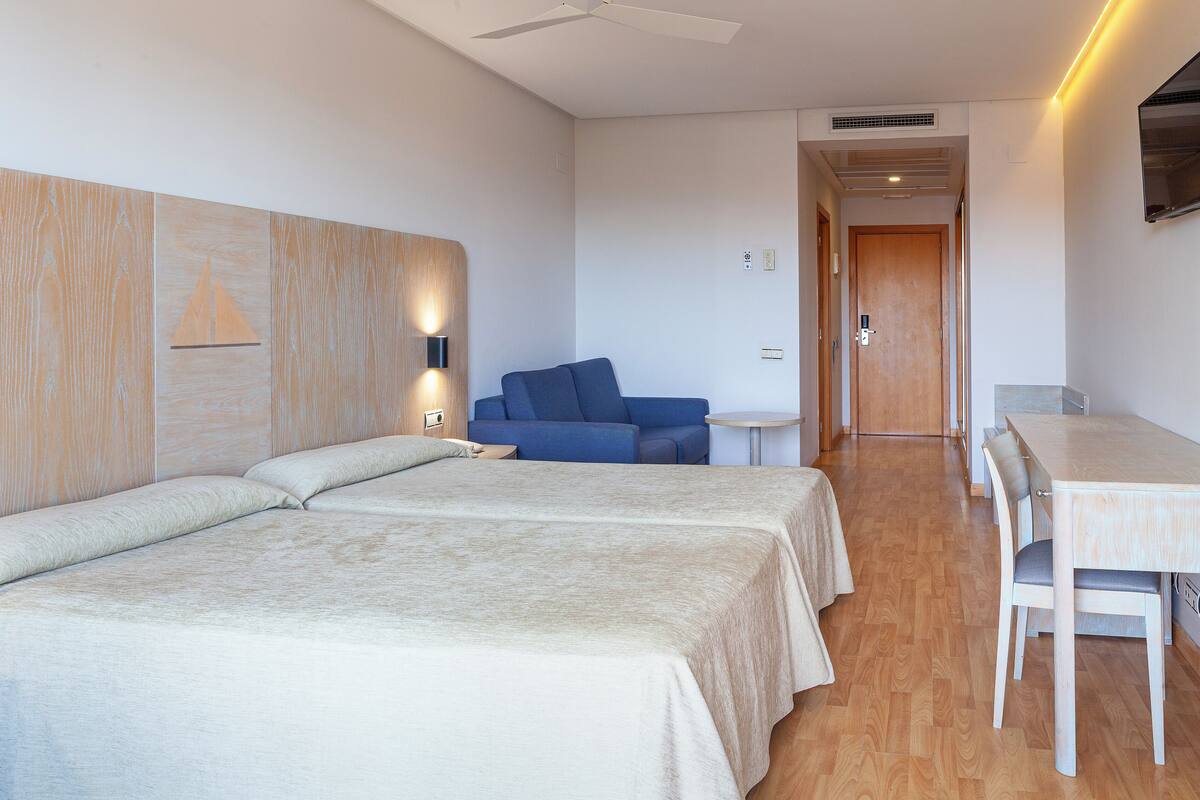 Ein Hotelzimmer mit zwei Betten, einem kleinen Tisch, einem Stuhl, einer Couch und einem Deckenventilator.