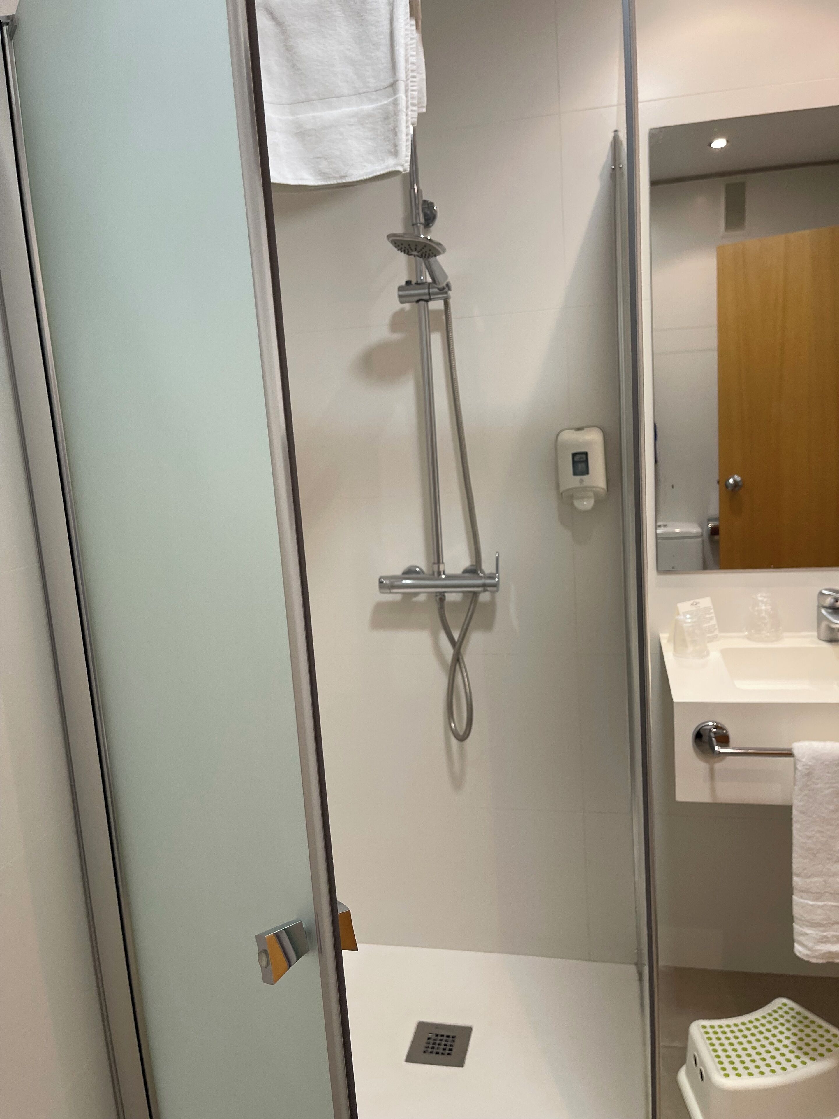 Un baño moderno con ducha envuelta en vidrio, un lavabo blanco con dispensador de jabón y una puerta de madera.
