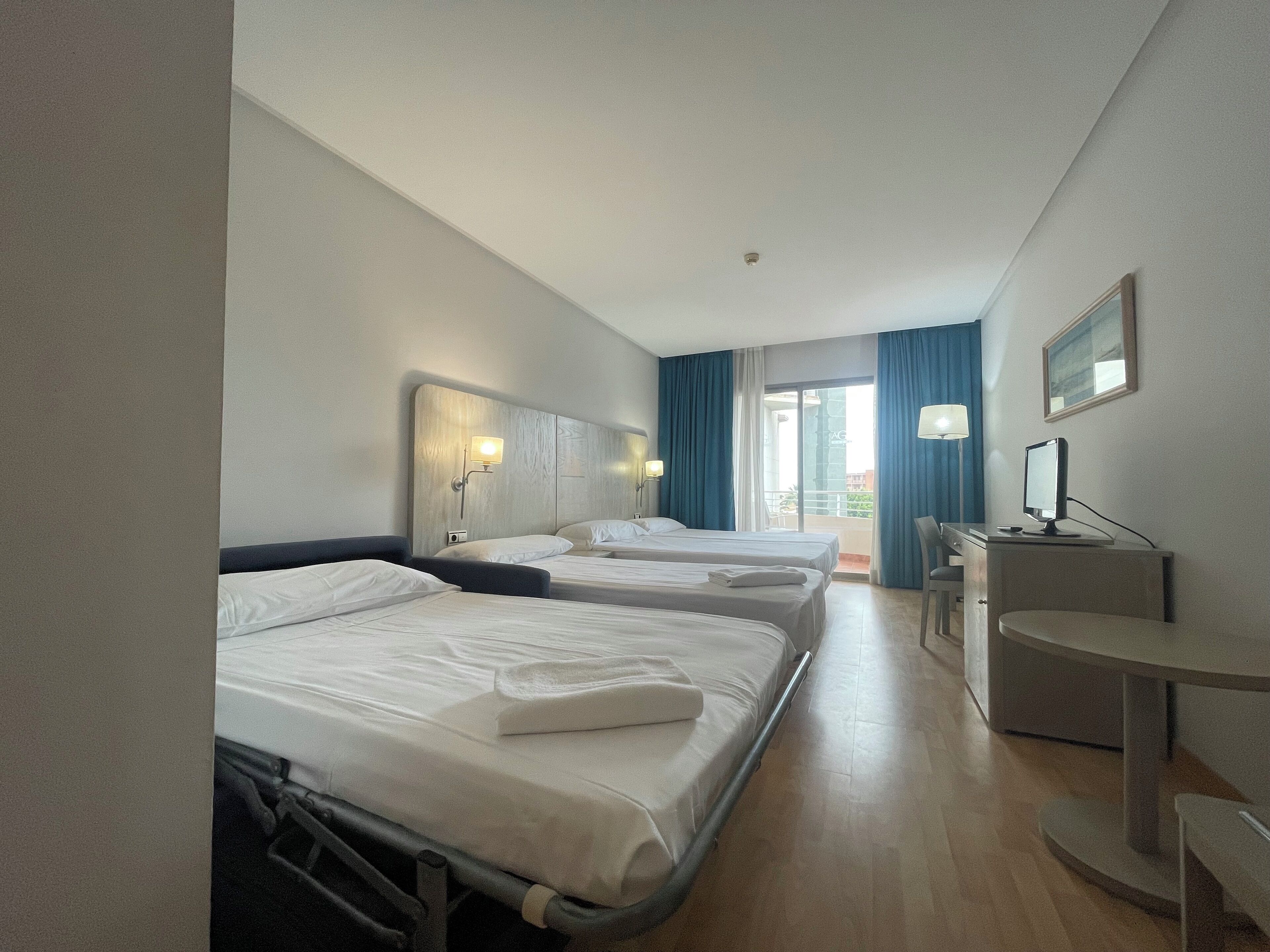 A hotel room with three beds, a desk with a computer, and a balcony with blue curtains.