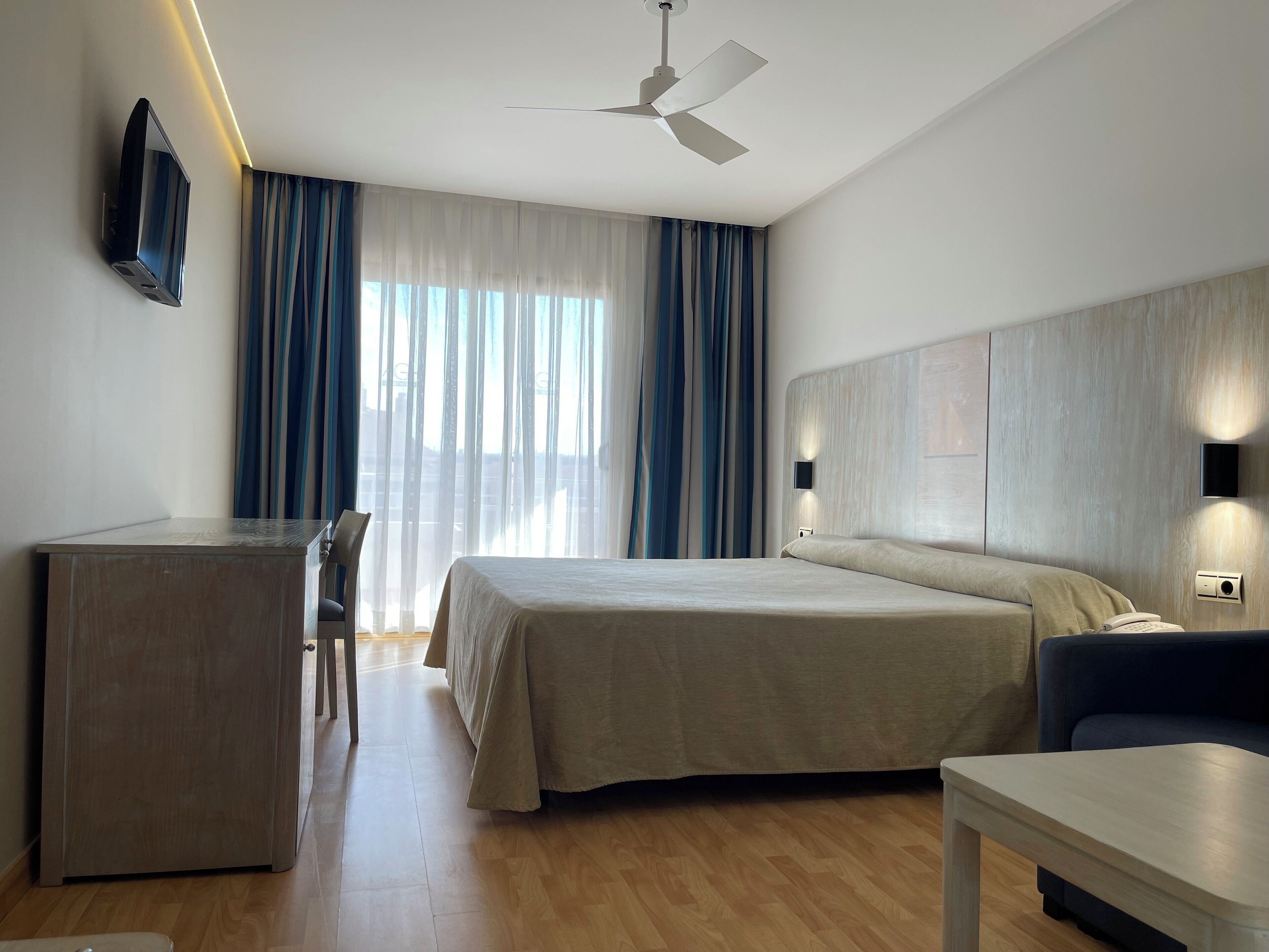 Habitación de hotel con cama, escritorio, silla, mesa, ventilador de techo y televisor montado en la pared.