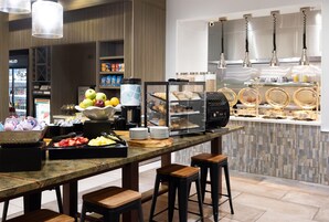 Un mostrador de desayuno con diversos alimentos, como frutas, pasteles y bebidas, en un entorno hotelero moderno.