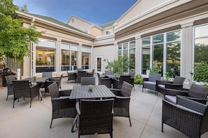 An outdoor patio area with multiple tables and chairs, large windows, and a tree. - Hilton Garden Inn Grand Forks - UND (Grand Forks)