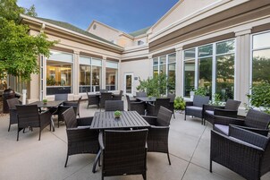 Un patio al aire libre con varias mesas y sillas, ventanas grandes y un árbol.