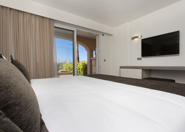 Una habitación de hotel moderna con una cama grande, un televisor de pantalla plana en la pared y un ventanal corredero que da a un balcón con vistas a zonas verdes y edificios.