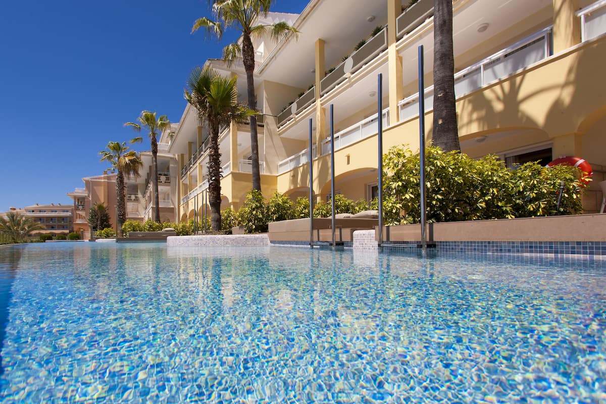 A swimming pool with clear blue water, surrounded by palm trees and a multi-story hotel building with balconies.