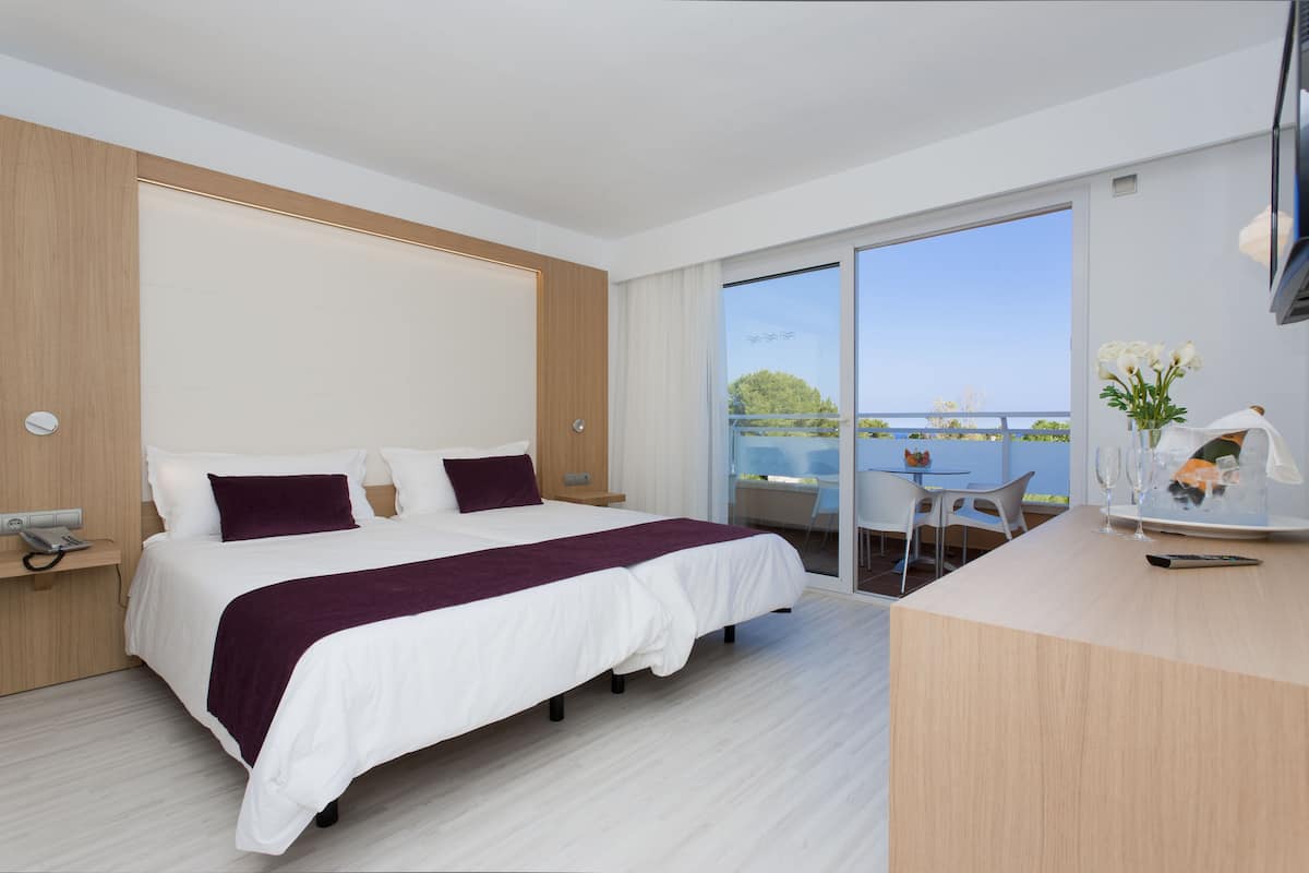 A modern hotel room with a large bed, a desk, and a balcony with a view of the sea.