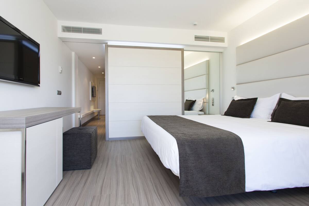 A modern hotel room with a bed, a TV mounted on the wall, a desk, and a wardrobe.