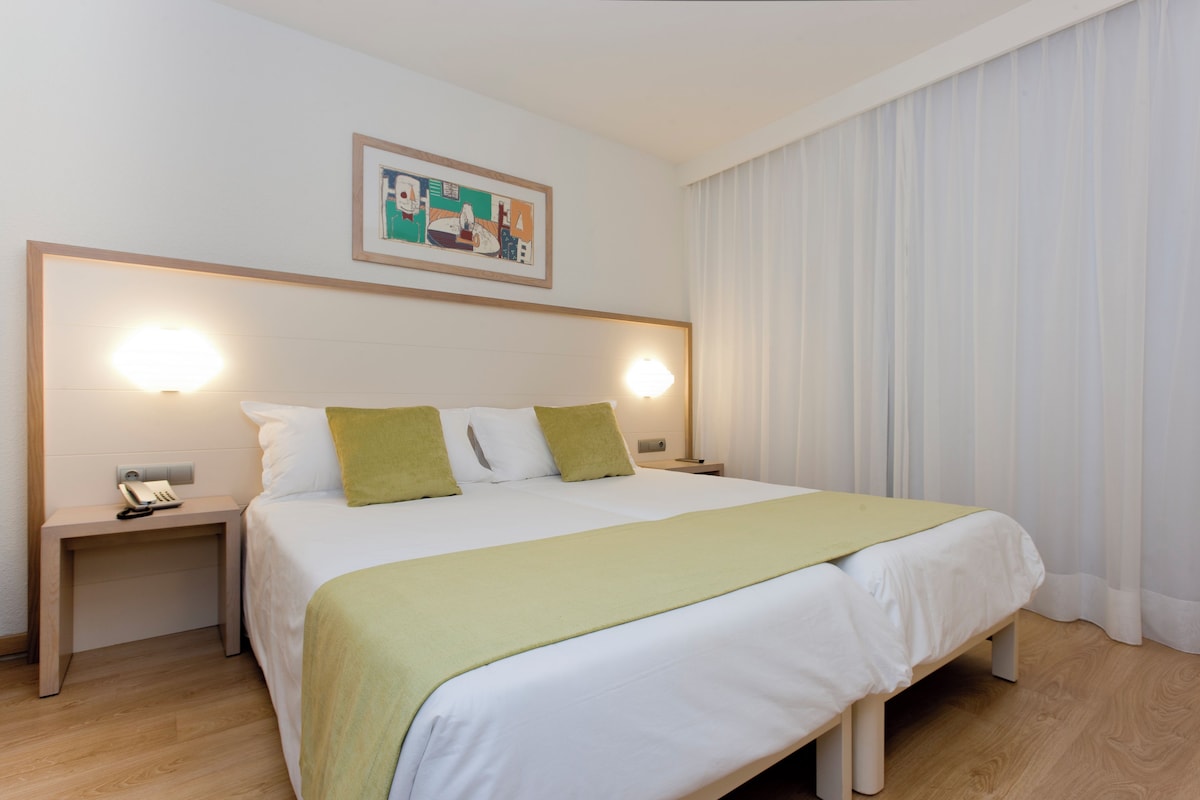 A neatly made bed with a green throw, two bedside lamps, a nightstand with a phone, and a painting above the bed.