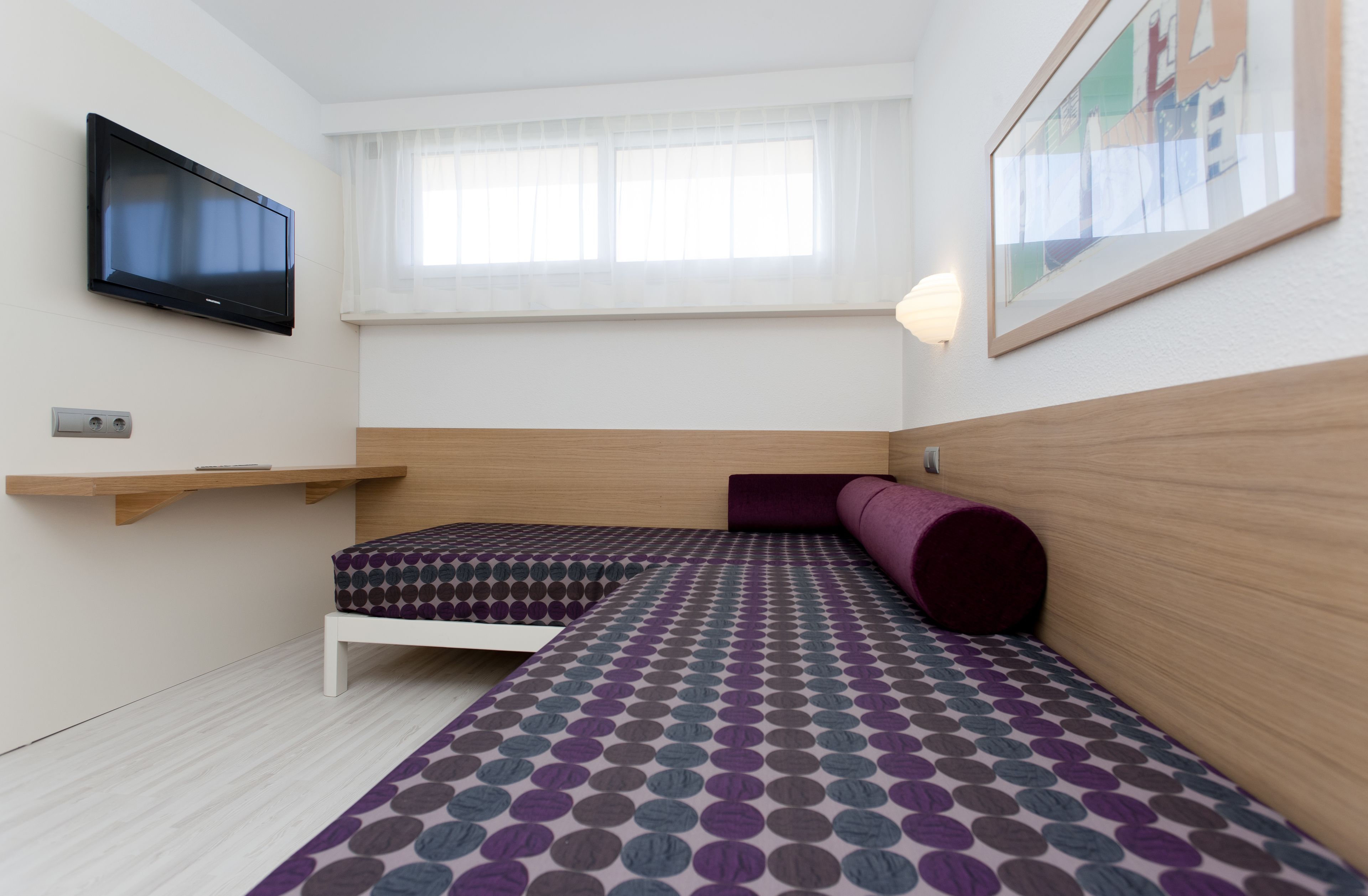 Una habitación pequeña y moderna con cama, televisor montado en la pared y ventana con cortinas translúcidas.