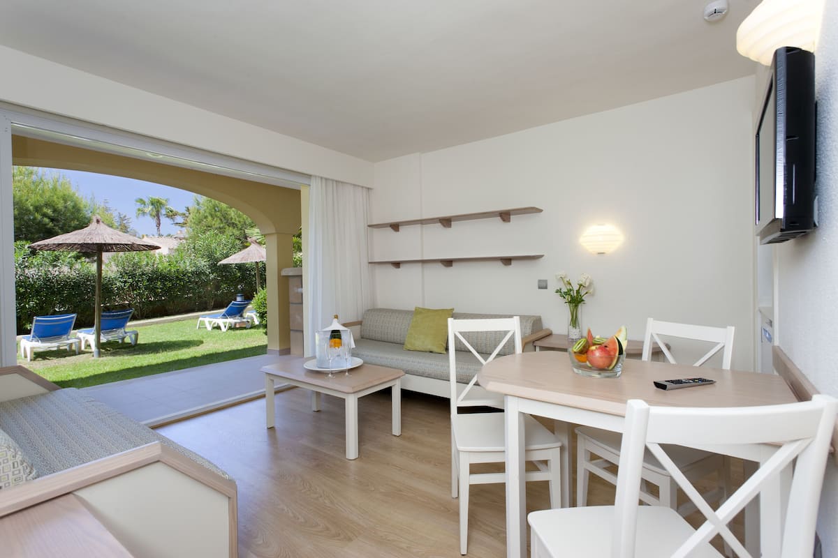A modern living room with a sofa, dining table, and chairs. There is a balcony with outdoor furniture and umbrellas.