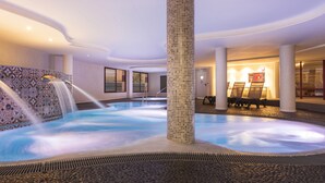 Una piscina interior con pared de mosaicos y una columna central, rodeada de asientos y vistas al exterior.