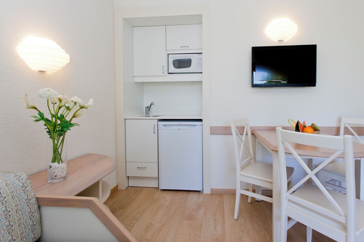 Una kitchenette compacta con microondas, pileta y heladera pequeña. Hay un comedor con mesa y sillas, y un jarrón decorativo con flores sobre un banco.