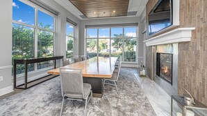 A spacious room with a large wooden table, multiple chairs, large windows, a fireplace, and a view of greenery outside.