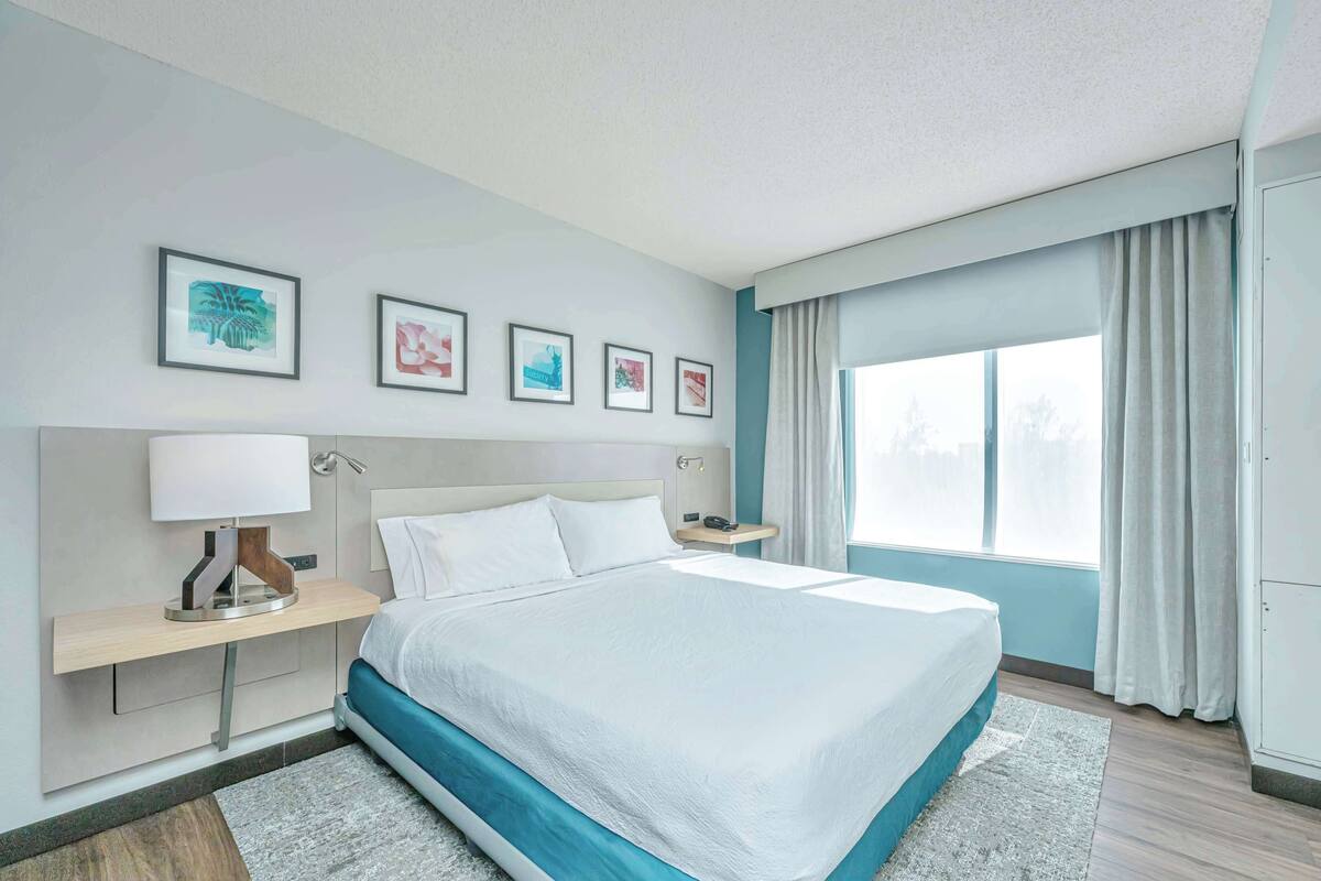 Une chambre d’hôtel moderne avec un grand lit, une table de chevet avec une lampe, une commode et une fenêtre avec des rideaux.
