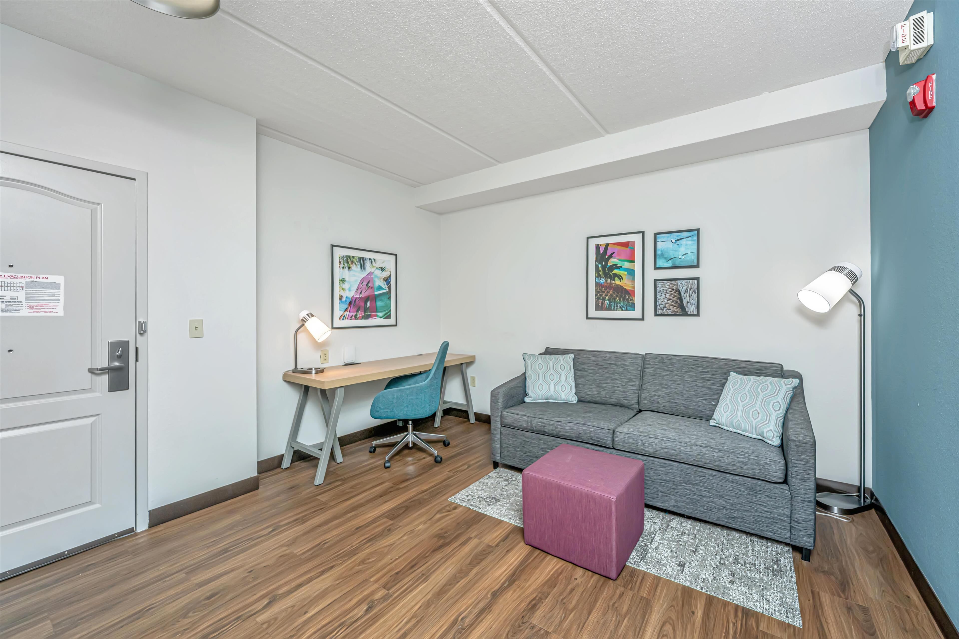 Une pièce moderne et bien éclairée, avec un plancher en bois, un canapé gris, un bureau avec une chaise et un petit pouf rose.