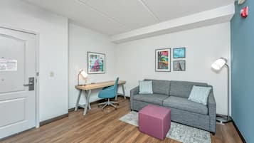 Une pièce moderne et bien éclairée, avec un plancher en bois, un canapé gris, un bureau avec une chaise et un petit pouf rose.