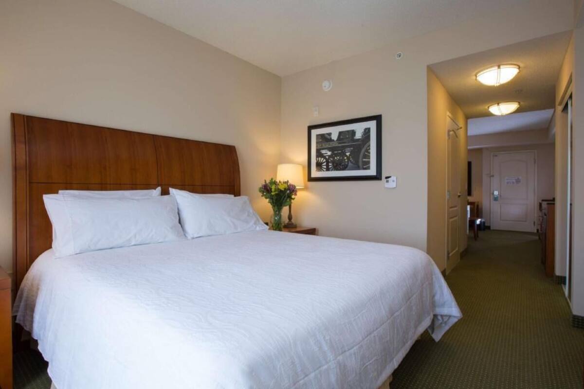 Une chambre d’hôtel avec un grand lit, une table de chevet avec une lampe, un tableau encadré au mur et un couloir menant à une porte.