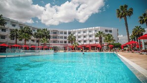Una piscina con sombrillas rojas, palmeras y un edificio de hotel al fondo.