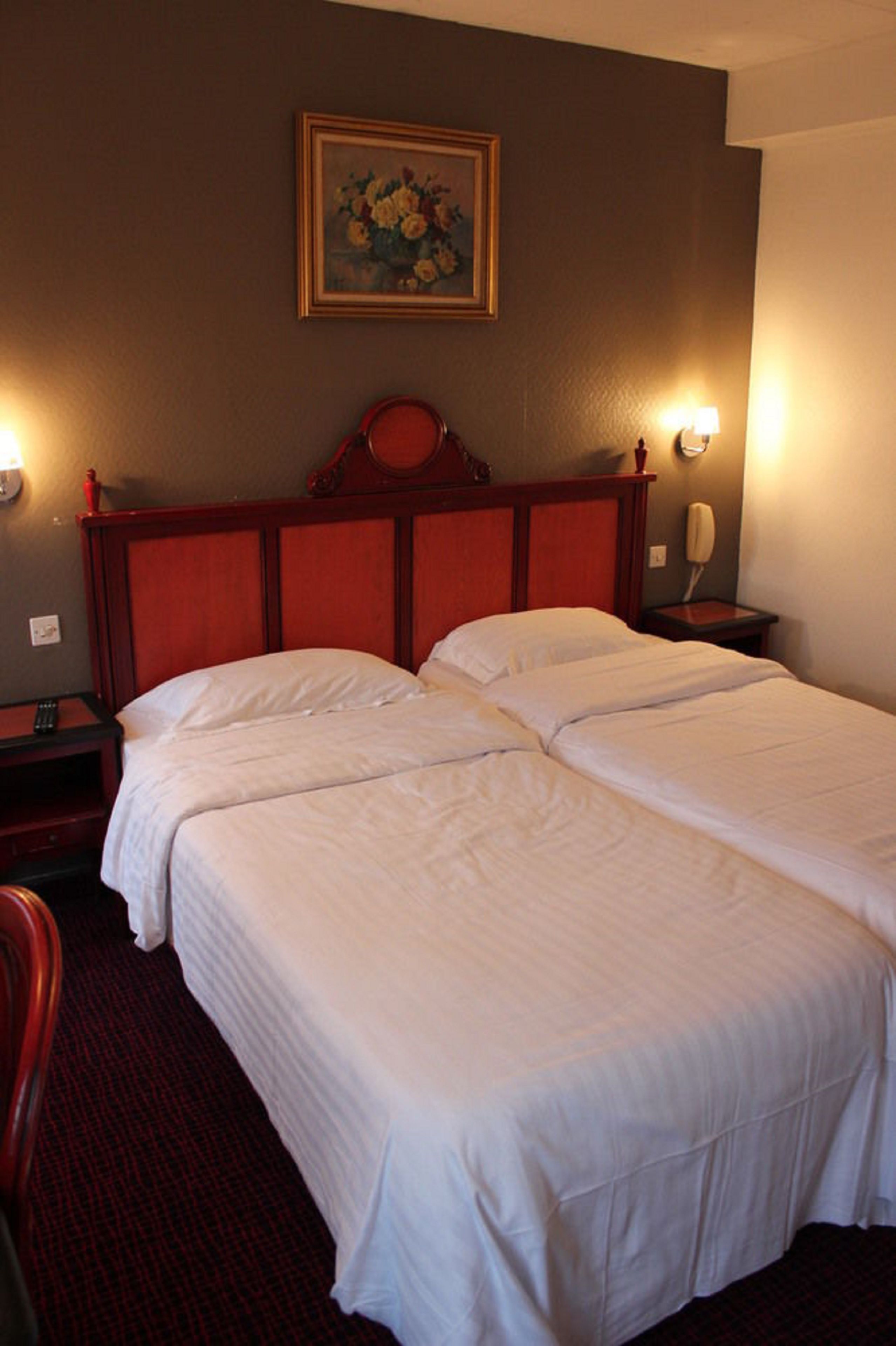 A hotel room with two single beds, a wooden headboard, a painting on the wall, and a bedside lamp.