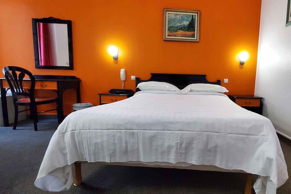 Quarto de hotel com uma cama, duas luminárias de cabeceira, uma escrivaninha com uma cadeira e um quadro na parede.