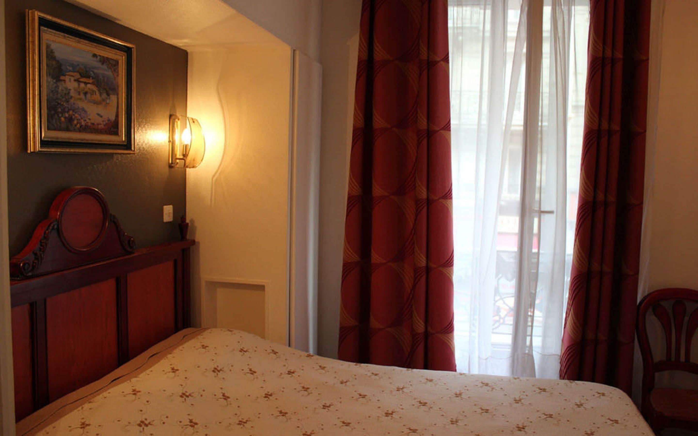 A hotel room with a bed, a wall-mounted light, a painting, and a window with curtains.