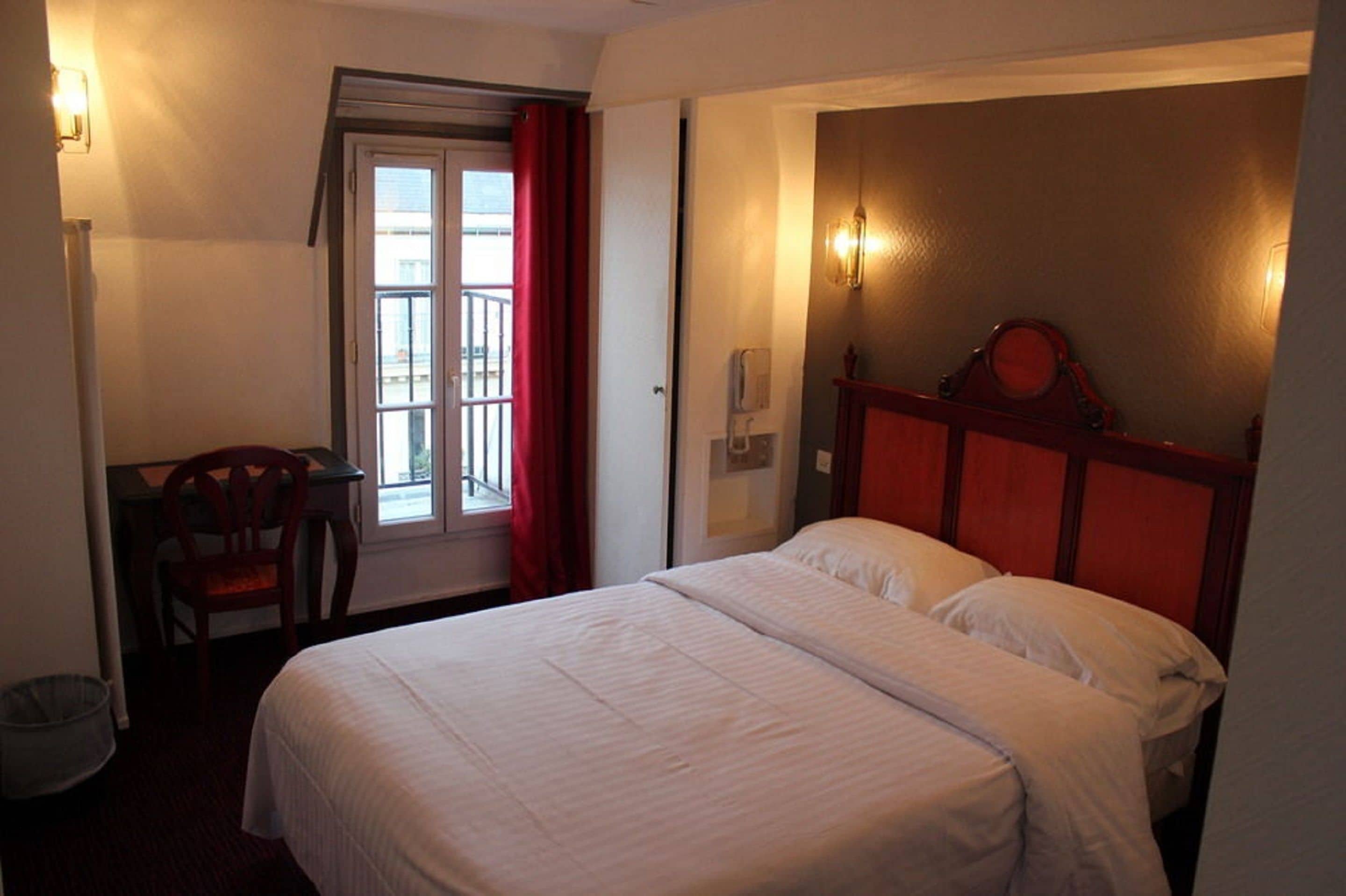 A hotel room with a bed, a chair, a desk, a window with red curtains, and a wall-mounted light fixture.