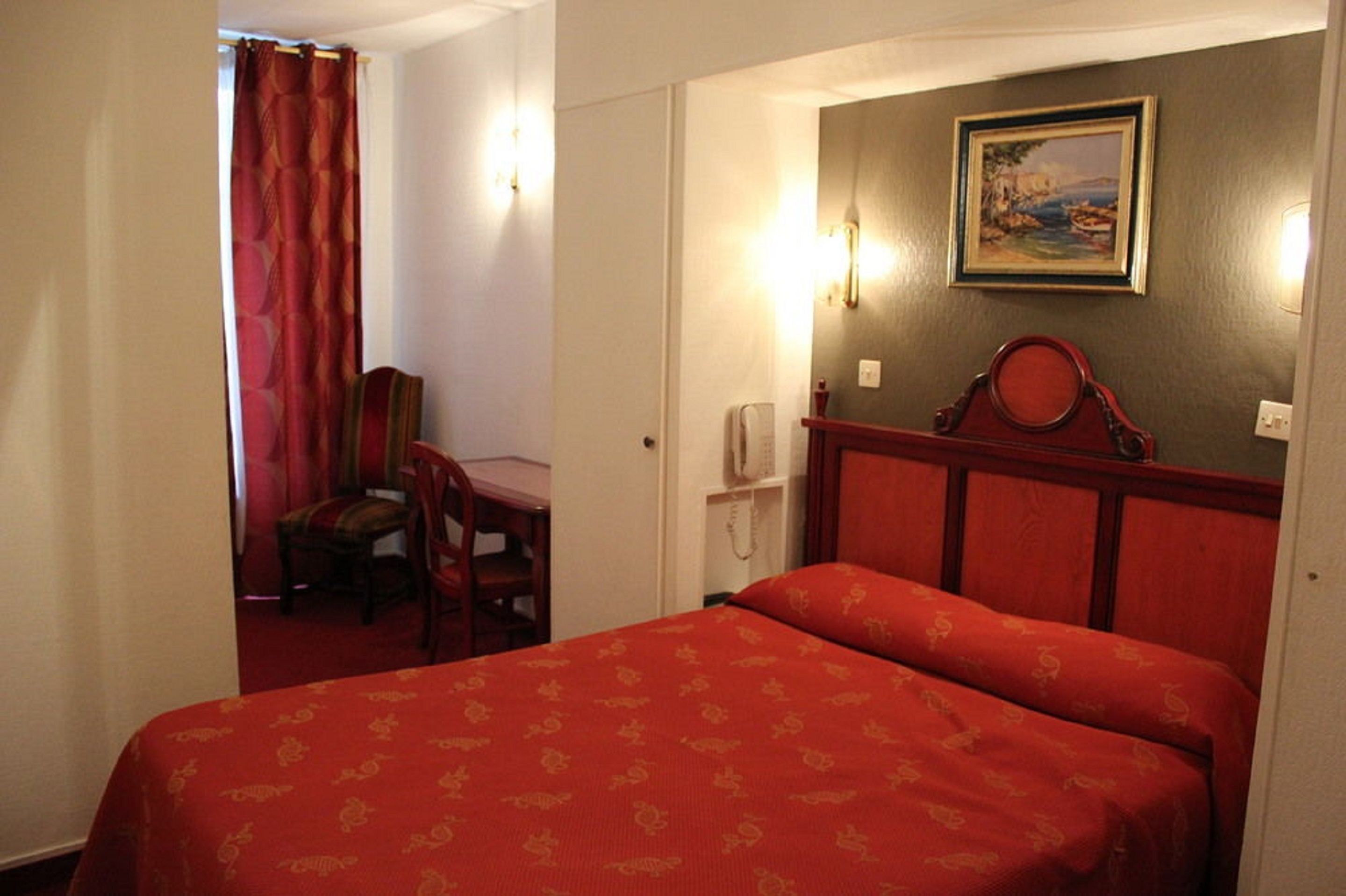 A hotel room with a bed, a chair, a desk, and a painting on the wall.