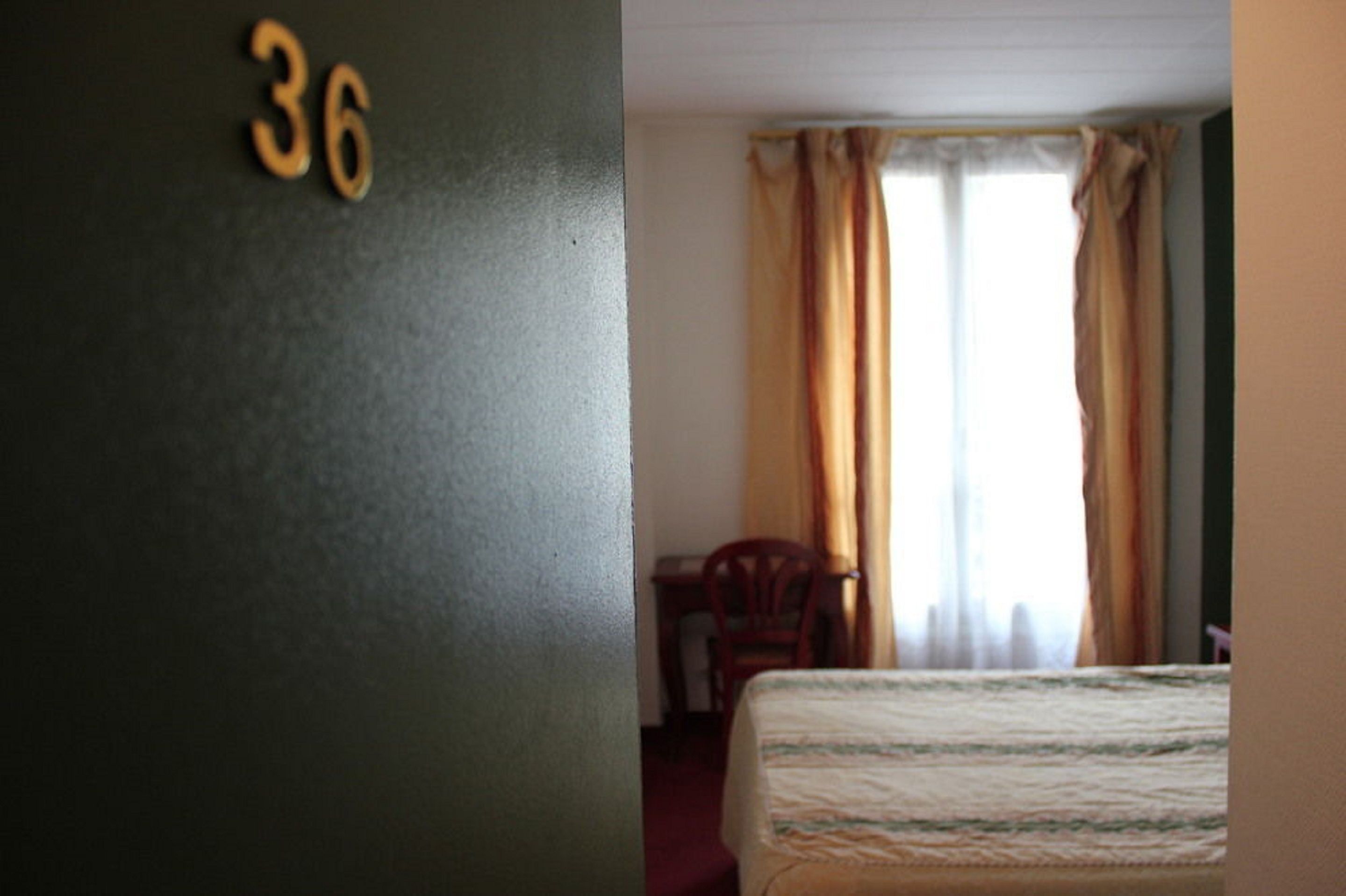 Una habitación con una cama, una silla y una ventana con cortinas. Se ve el número 36 en la pared.
