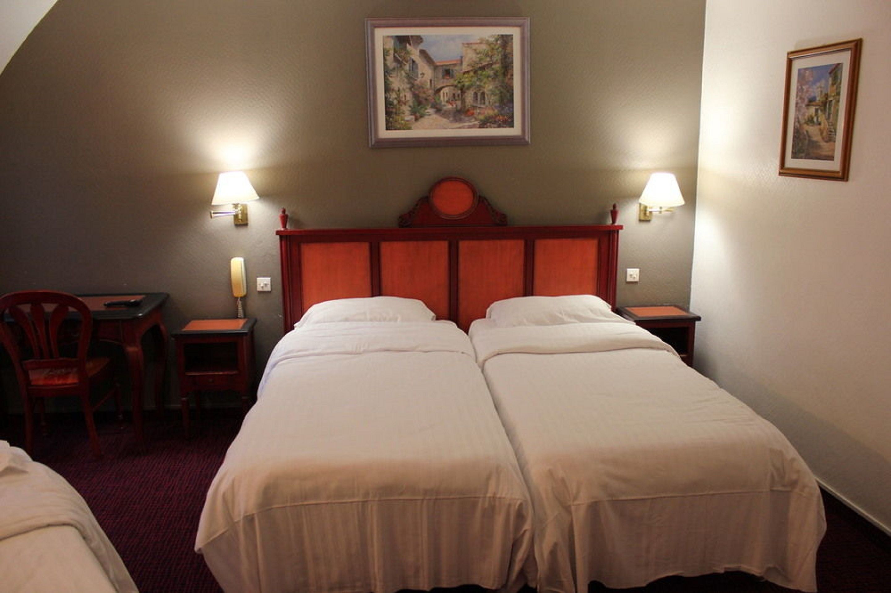 A hotel room with two single beds, a wooden headboard, two bedside lamps, a desk with a chair, and a framed painting on the wall.