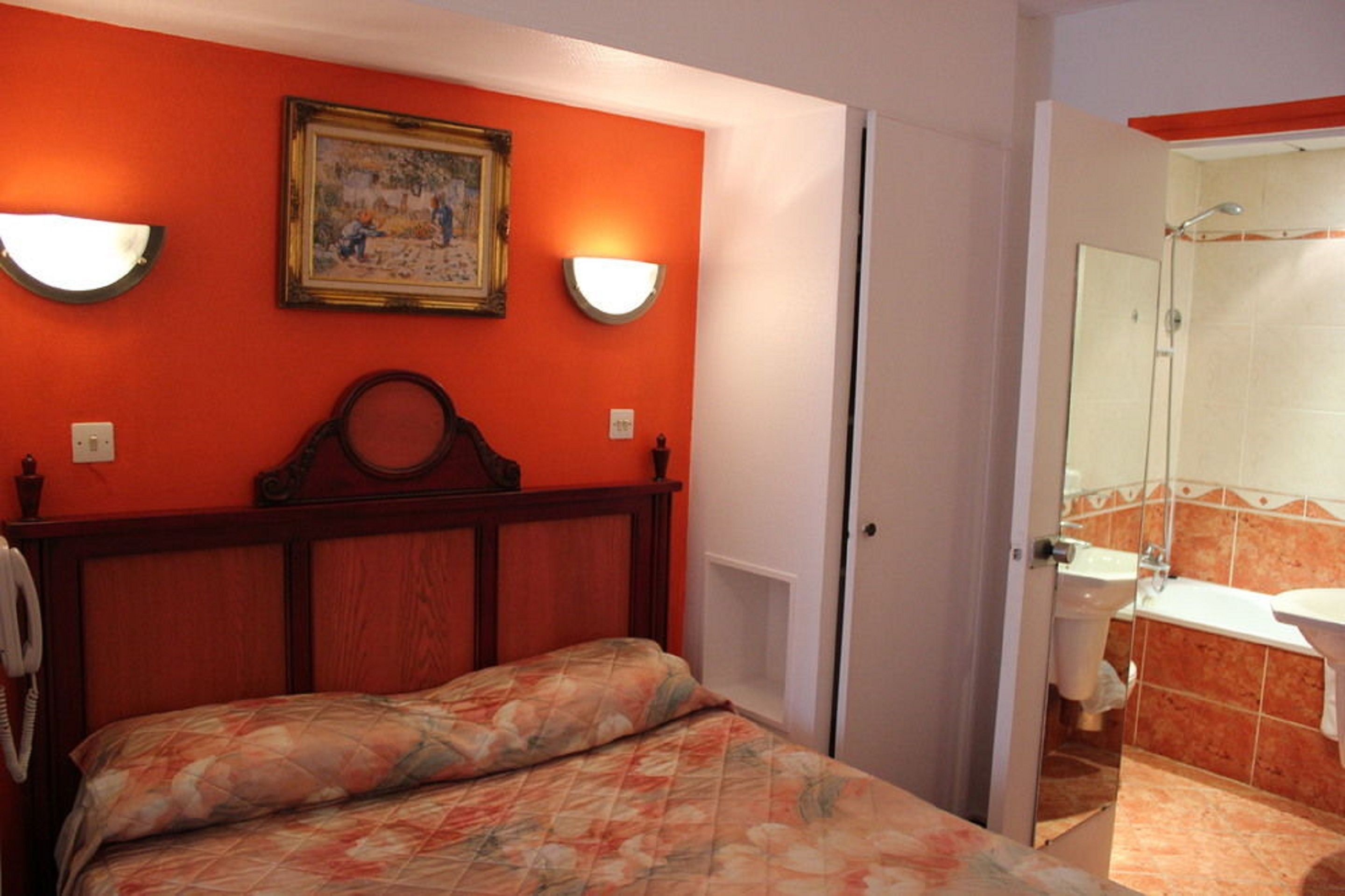 A bedroom with a wooden headboard, a bed with floral bedding, two wall-mounted lights, and a painting above the headboard.