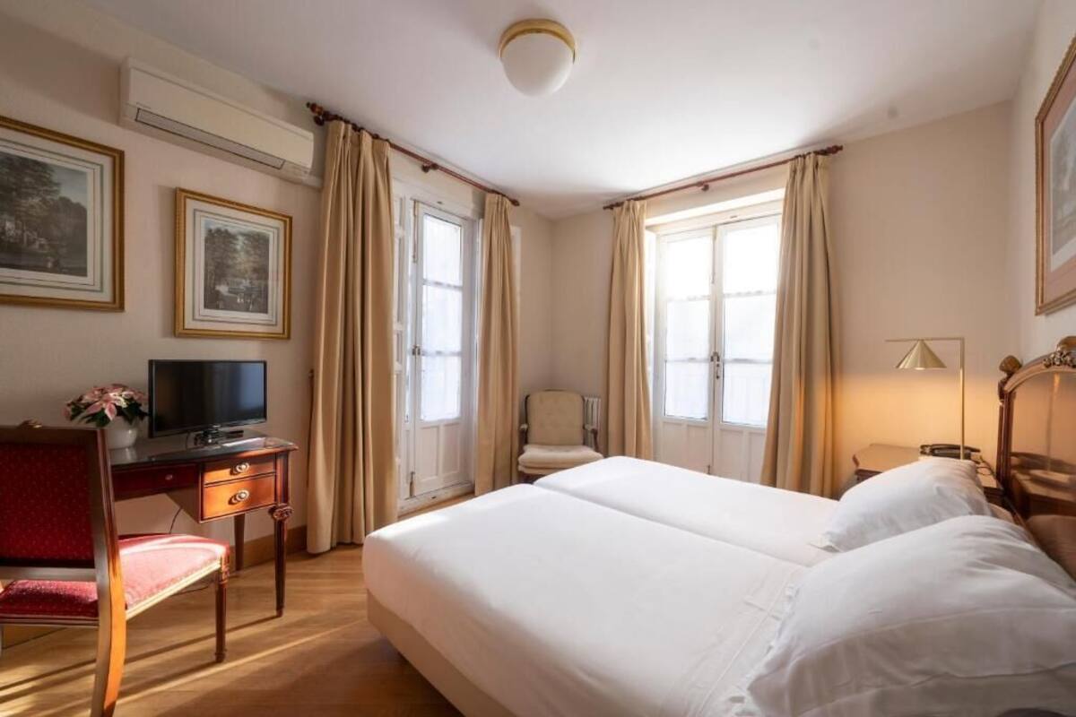 Una habitación de hotel con cama, escritorio con televisor, una silla y dos ventanas con cortinas.