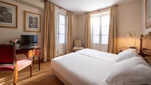 Una habitación de hotel con cama, escritorio con televisor, una silla y dos ventanas con cortinas.