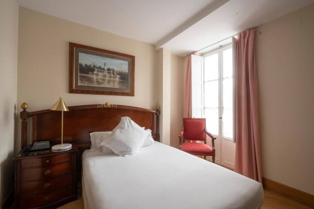 Un cuarto de hotel con cama, una mesita de noche con lámpara, una silla, una ventana con cortinas y un cuadro en la pared.