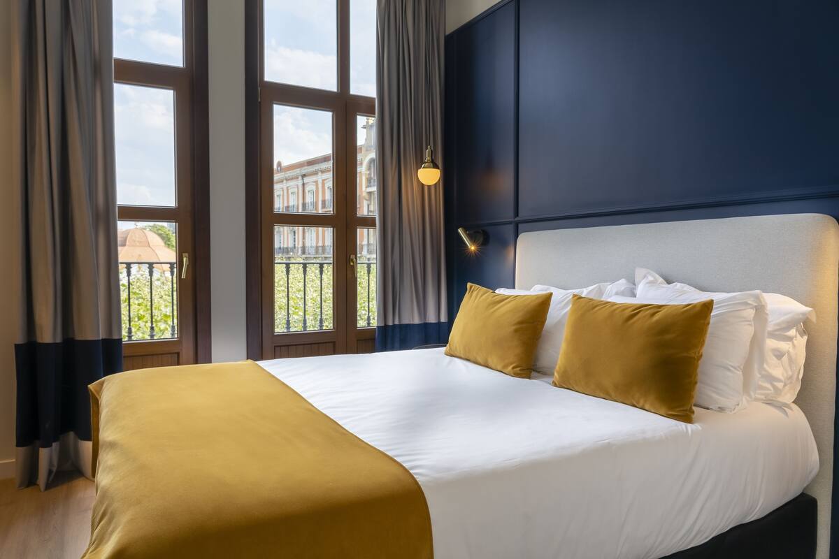 A hotel room with a large bed, a view of a building through the window, and a nightstand with a lamp.