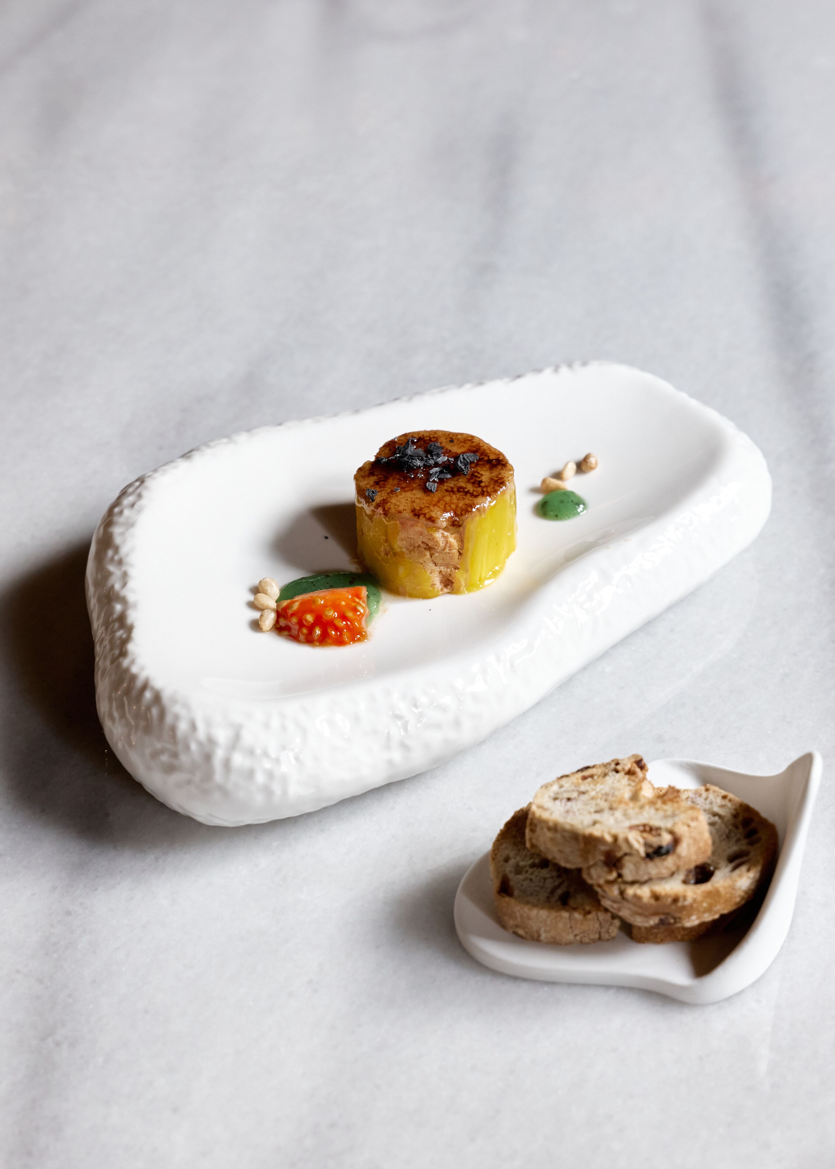 Un dessert servi dans une assiette en céramique blanche à bords texturés, accompagné d’un petit panier de pain.