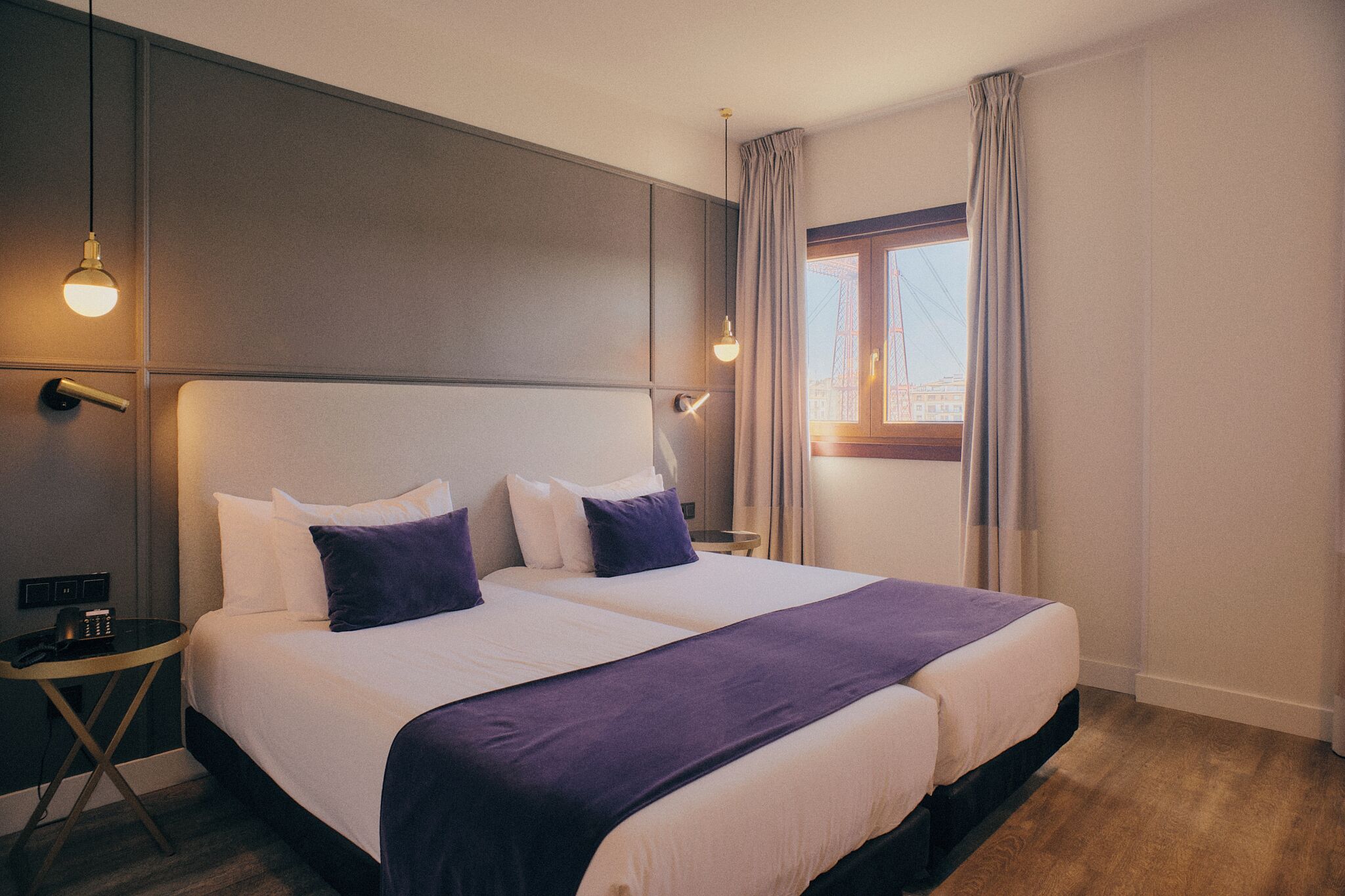 Una cama doble con ropa de cama blanca y morada, una mesita de noche con un teléfono y una ventana con cortinas.