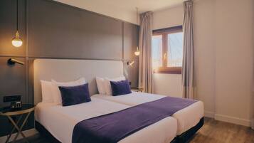 Una cama doble con ropa de cama blanca y morada, una mesita de noche con un teléfono y una ventana con cortinas.