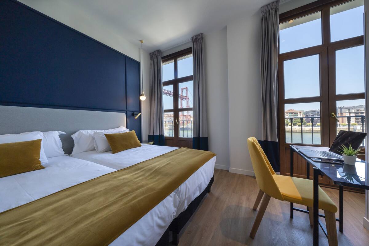 A hotel room with two beds, a desk with a laptop, a chair, and large windows with a view of buildings and cranes.