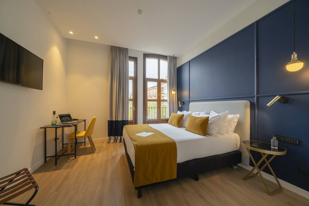 A modern hotel room with a bed, a desk with a laptop, a chair, and a small table.