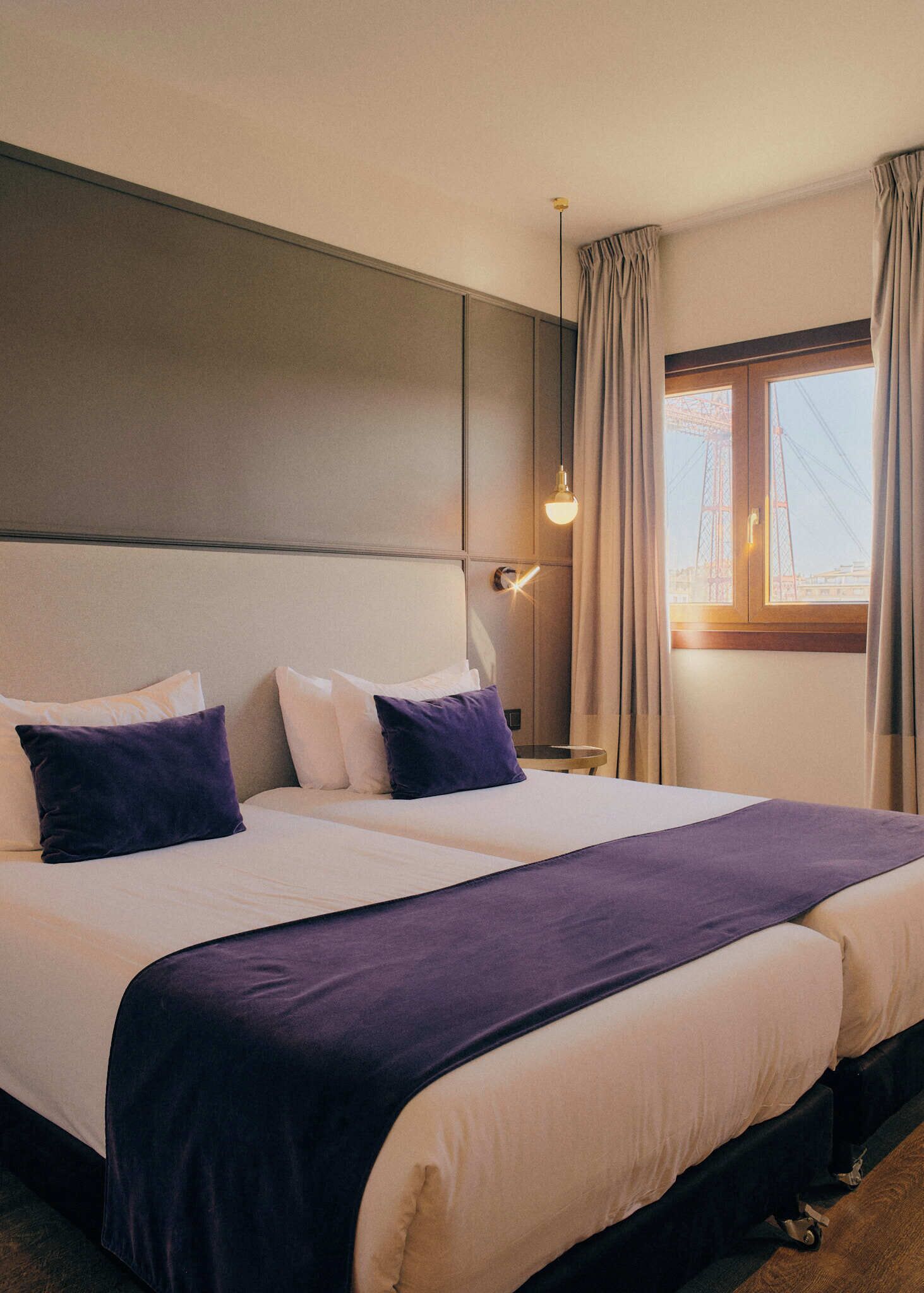 A hotel room with a large bed, two pillows, a bedside table, a lamp, and a window with curtains.