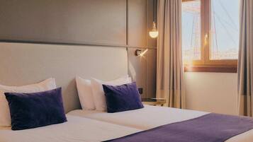 Habitación de hotel con una cama grande, dos almohadas, una mesita de noche, una lámpara y un ventanal con cortinas.