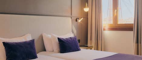 A hotel room with a large bed, two pillows, a bedside table, a lamp, and a window with curtains.