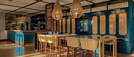 Zona de bar con taburetes de madera, lámparas colgantes y una pared azul con estantes llenos de botellas.
