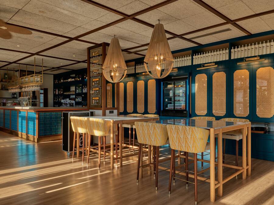Zona de bar con taburetes de madera, lámparas colgantes y una pared azul con estantes llenos de botellas.